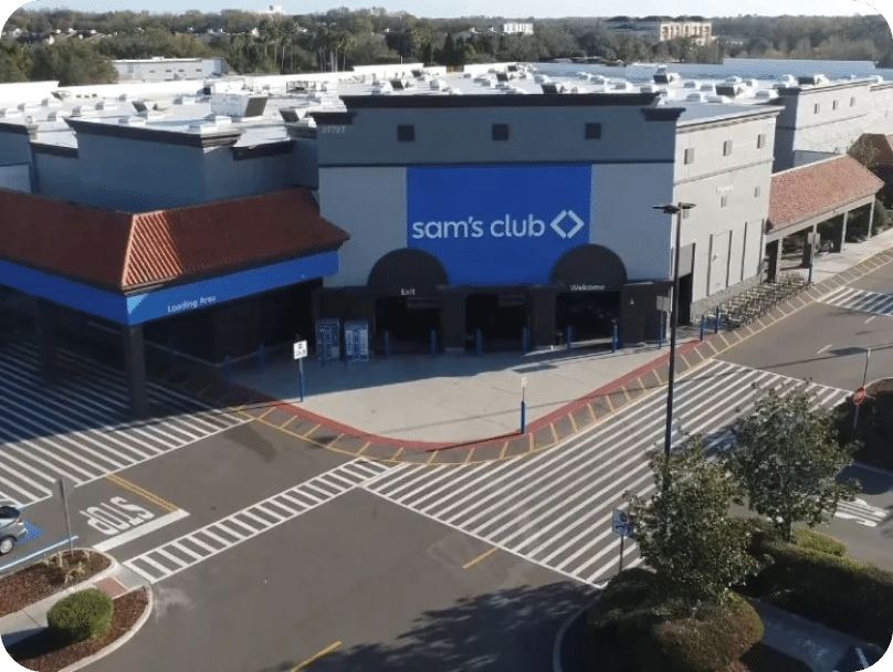 Aerial view of the entrance to a Sam's Club store. The building features a blue and gray facade with the Sam's Club logo. There are parking spaces and shopping carts visible in the foreground, and the area appears well-maintained with some greenery.