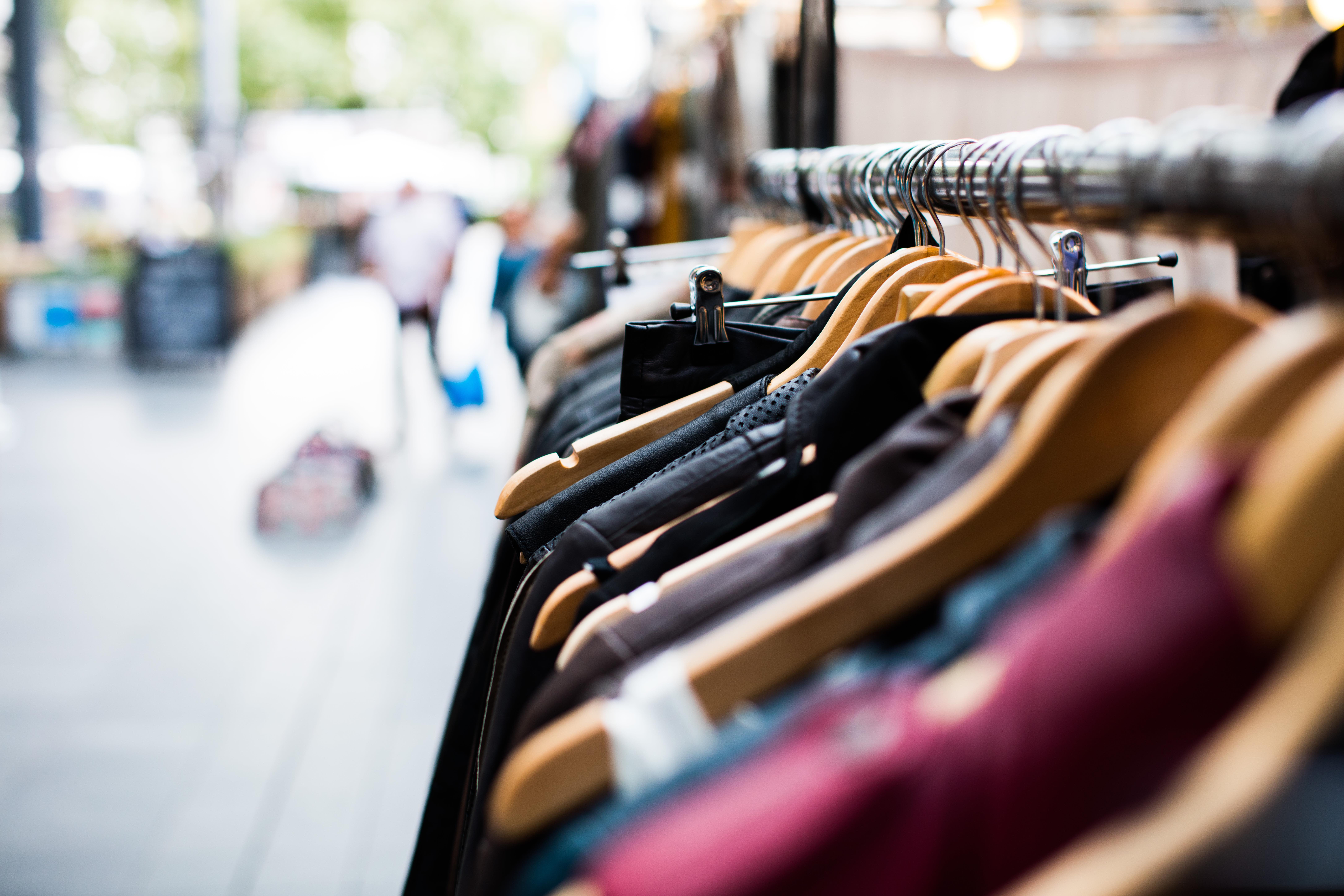 photo-1470309864661-68328b2cd0a5-copy Close-up view of clothing items hanging on a rack with wooden hangers. The background is blurry, with people walking and a bright, outdoor market setting. The clothes include various colors and styles.