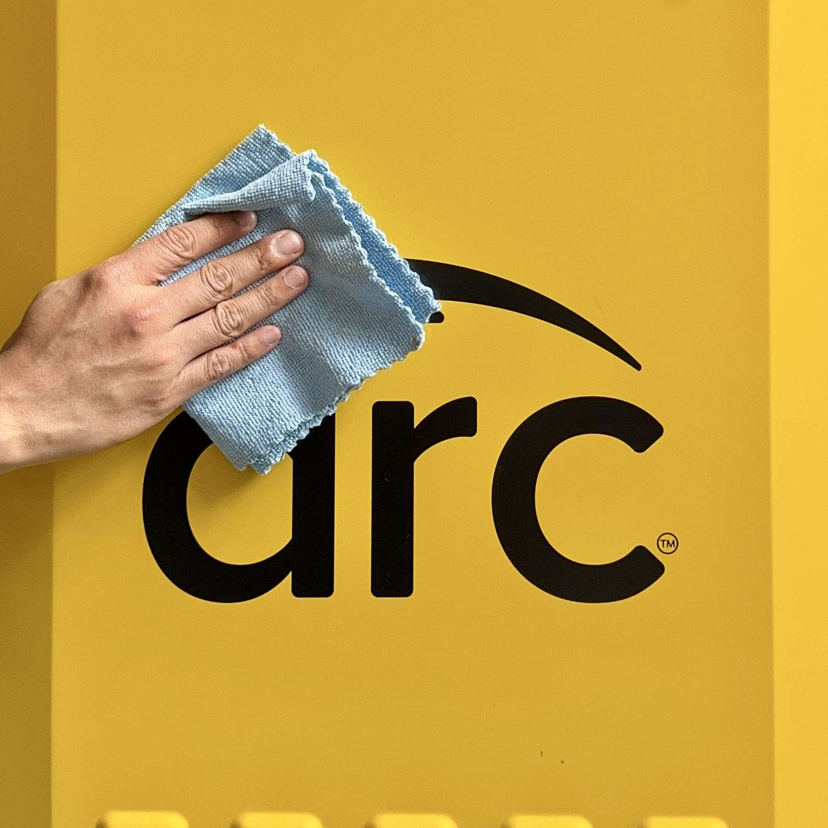 A hand is cleaning a black logo that reads "arc" on a yellow background using a blue cloth. The "C" in "arc" has a curved line over it. The background is uniformly yellow.