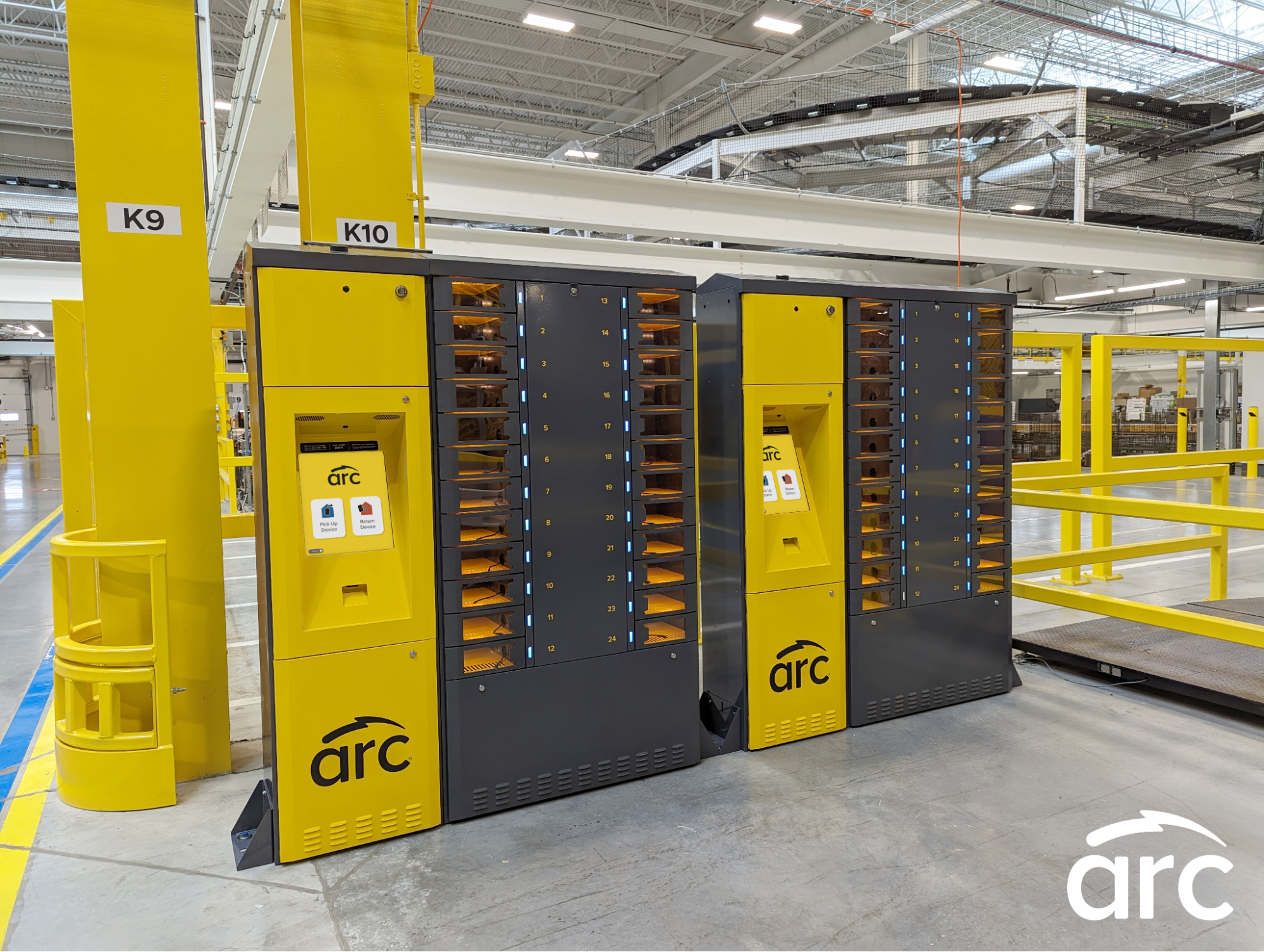 A warehouse worker in an orange safety vest and gray t-shirt is looking at a smartphone. The worker is standing in front of red metal shelving units filled with cardboard boxes and various items. The background shows a typical warehouse environment.